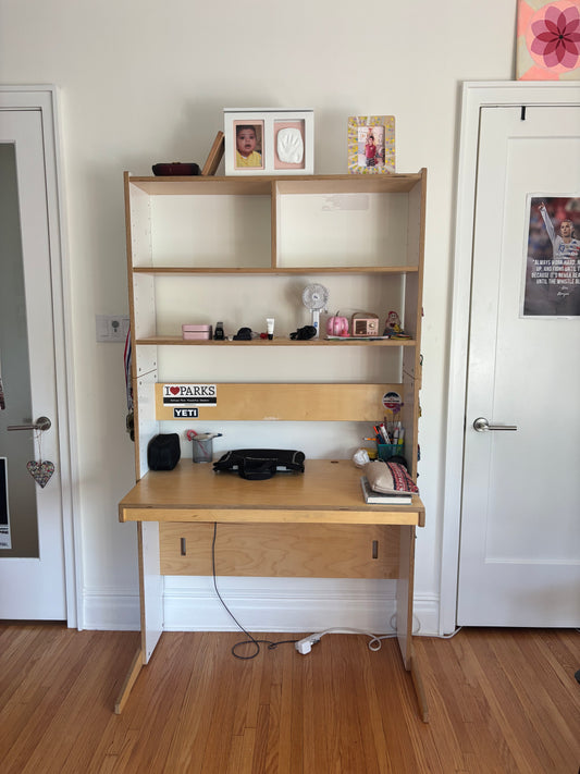 A light wood vanity or desk organizer with multiple open shelves and compartments, shown in a real room with white walls, a wood floor, and household items inside.