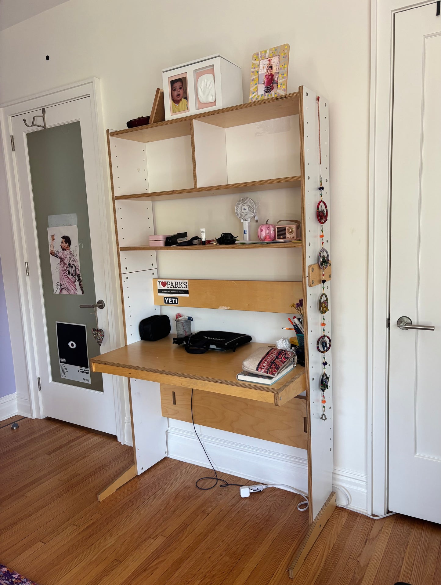A light wood desk with an attached open shelving unit, shown in a home office setting with wood floors, white walls, and two doors.