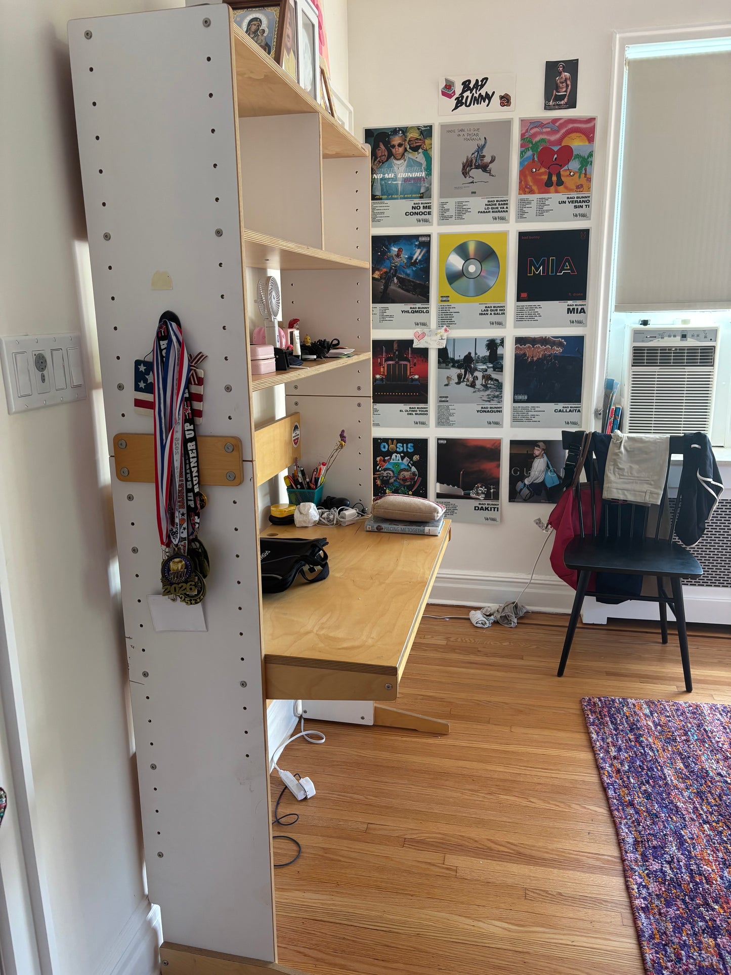 A tall white pegboard wall organizer with open shelves and a desk attachment in a bedroom. Medals hang from a side rack, and the wall behind is covered with music posters and a chair is visible in the room.