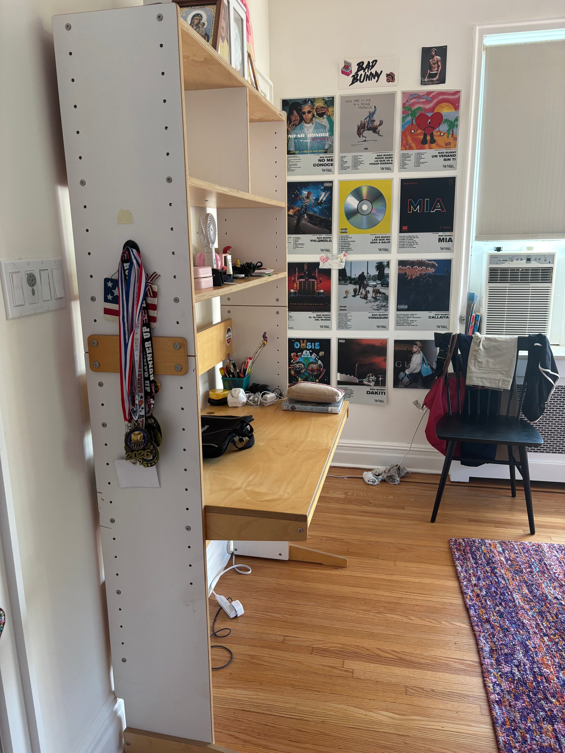 A tall white pegboard wall organizer with open shelves and a desk attachment in a bedroom. Medals hang from a side rack, and the wall behind is covered with music posters and a chair is visible in the room.