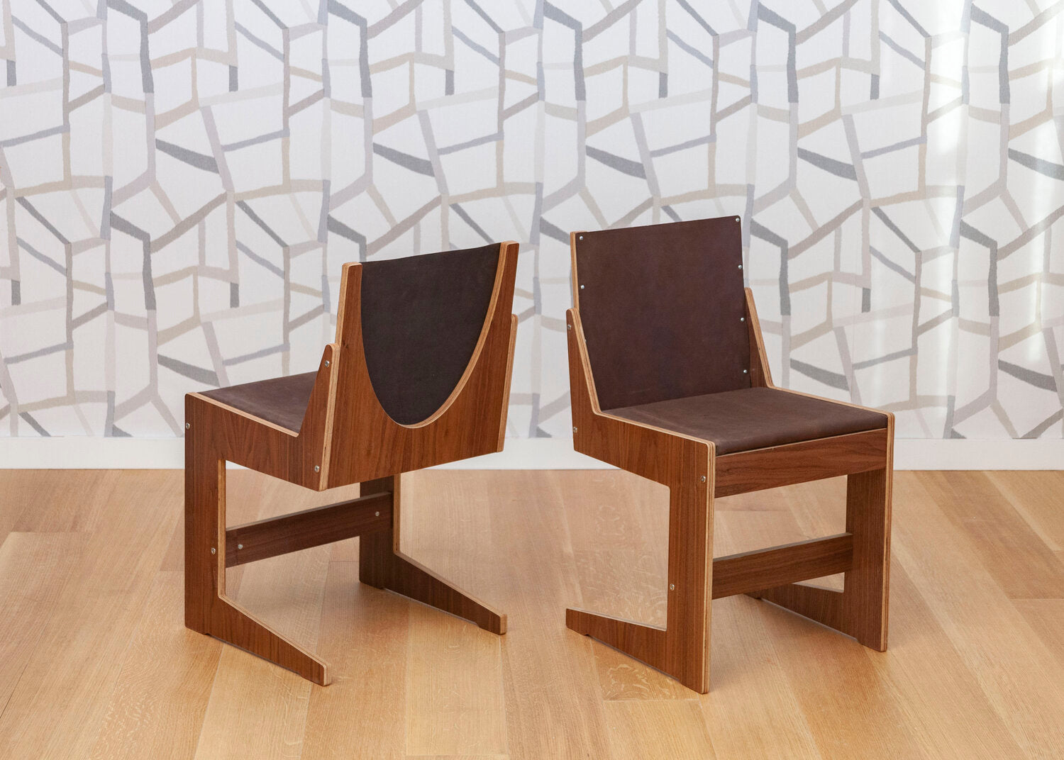 Two contemporary wooden chairs with black leather backrests on a hardwood floor against a geometric wallpaper.