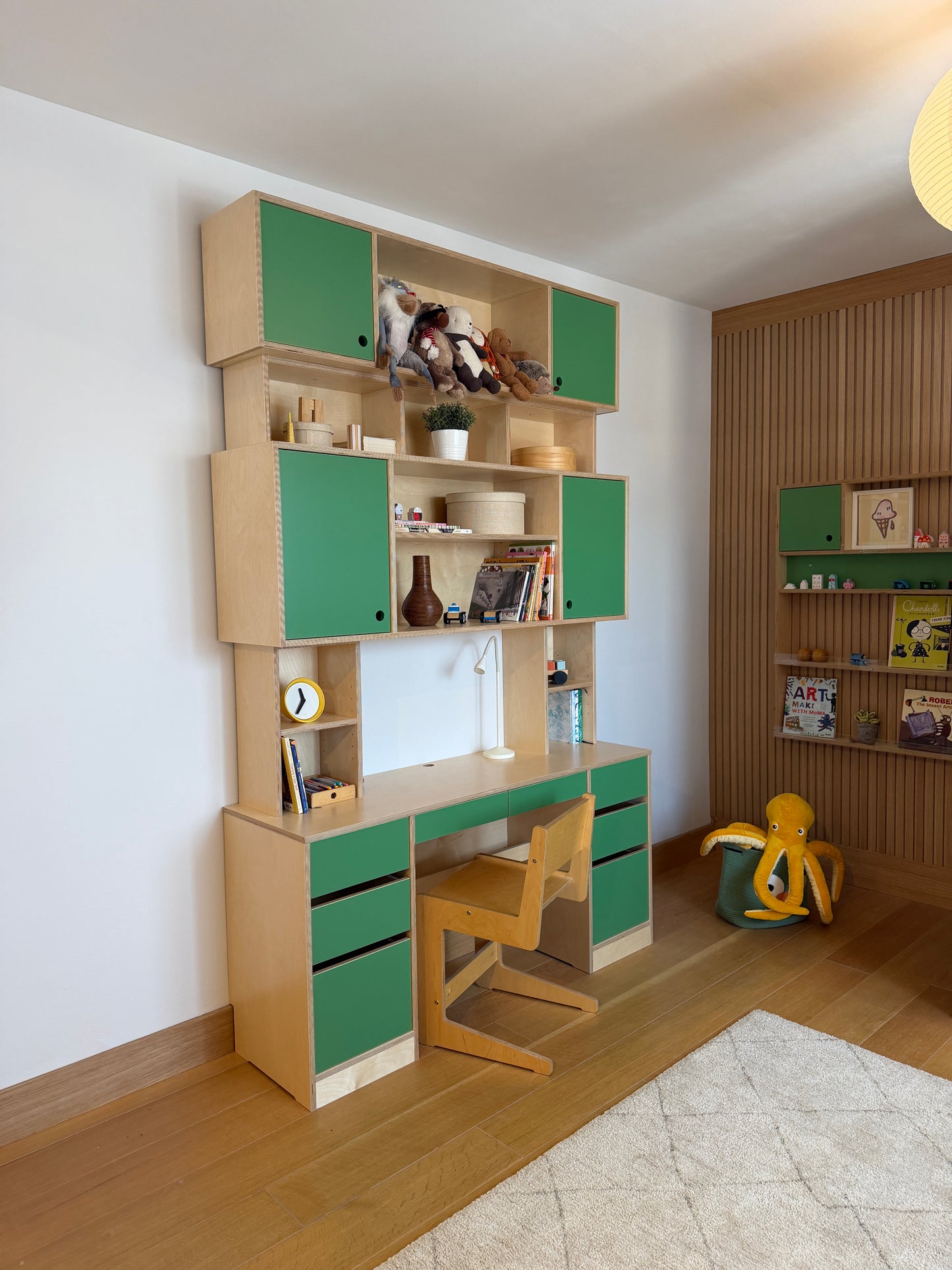 A modular wooden children's play kitchen or activity room set with green-leaf style lids, open compartments, toy storage, and two wooden chairs, shown from an elevated angle in a room with wood flooring and wall shelving.