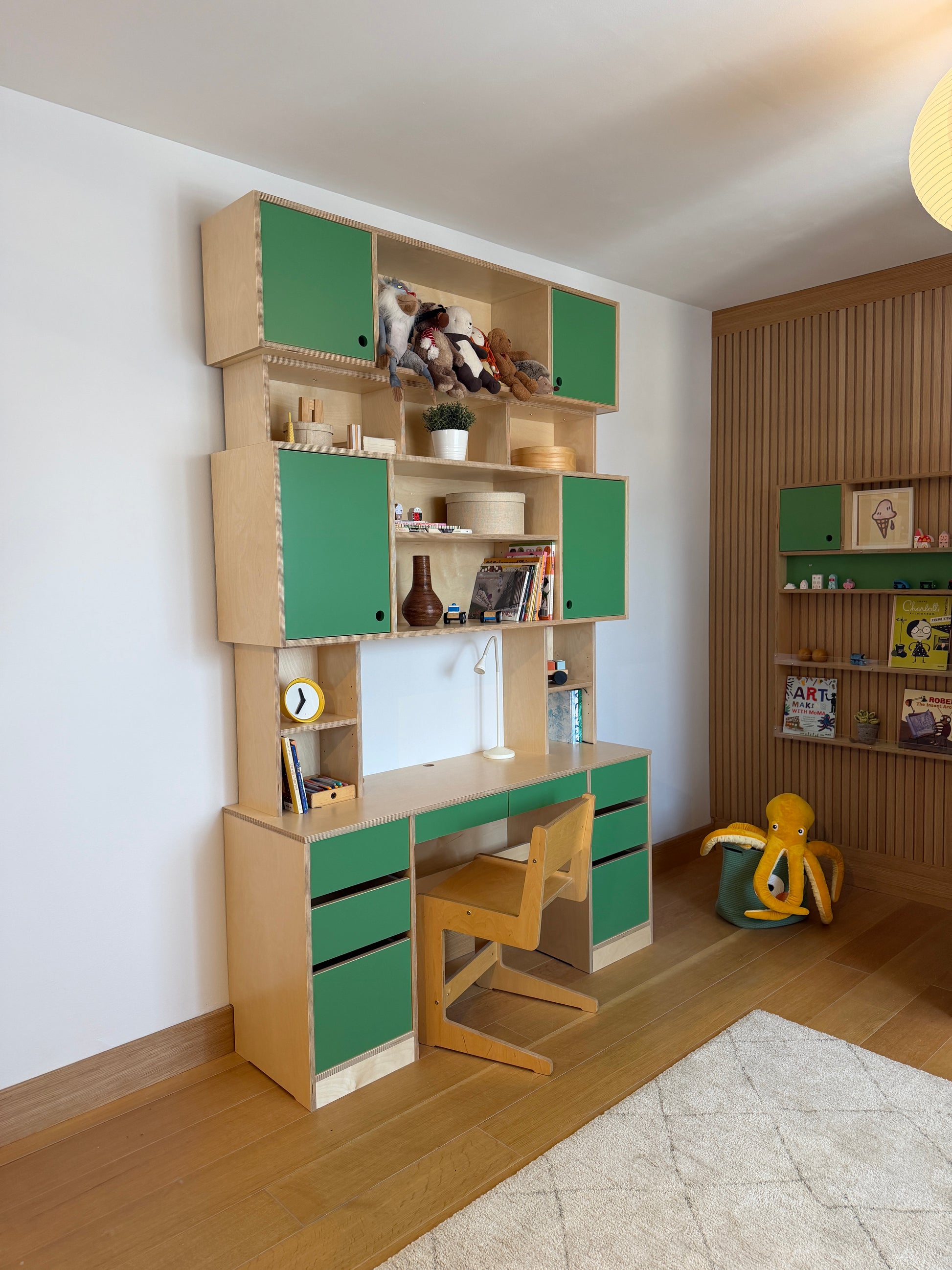 A modular wooden children's play kitchen or activity room set with green-leaf style lids, open compartments, toy storage, and two wooden chairs, shown from an elevated angle in a room with wood flooring and wall shelving.