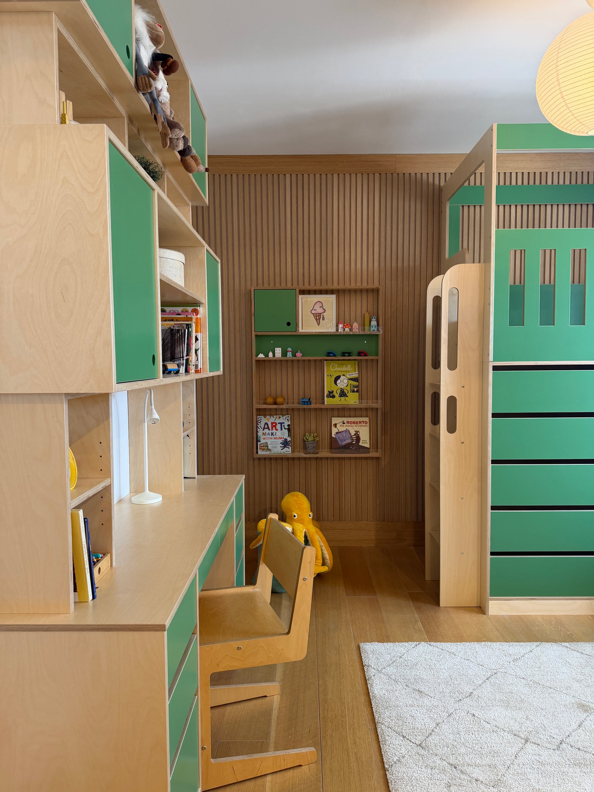 A children's bedroom featuring a light wood and mint green modular furniture set, including a desk, chair, bookshelves, storage cabinets, and a loft bed frame, with a slatted wood accent wall behind.