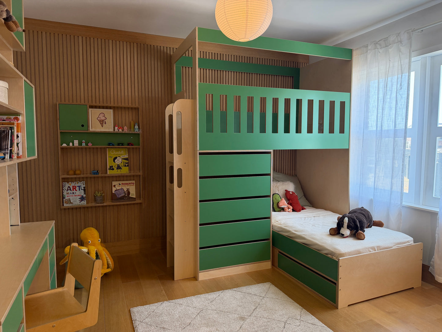 A children's bedroom featuring a green and light wood bunk bed with an attached storage cabinet, desk, and shelving against a slatted wood wall.