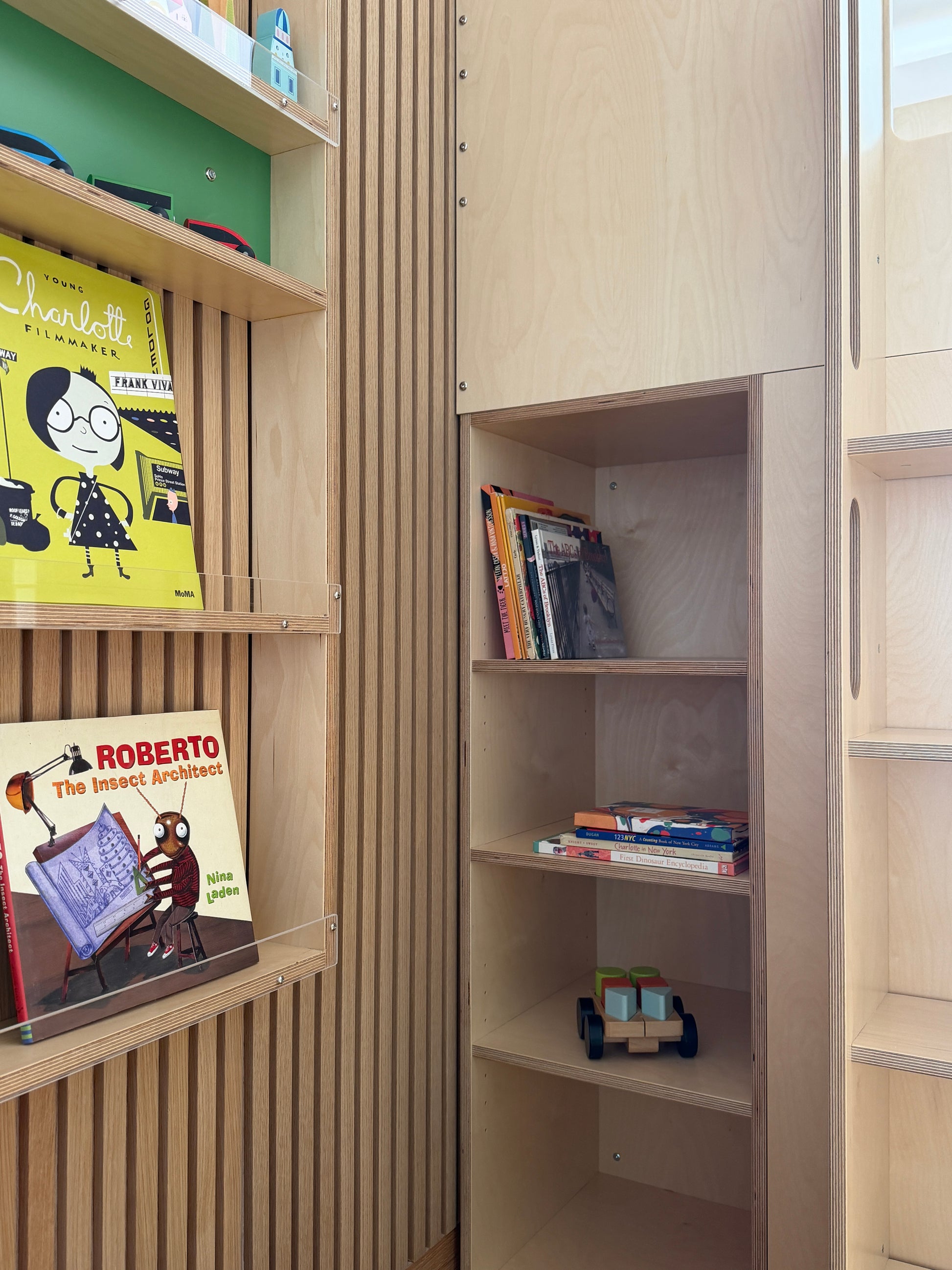 Light natural wood children's bookshelves with slatted paneling, holding colorful children's books and a small toy car.