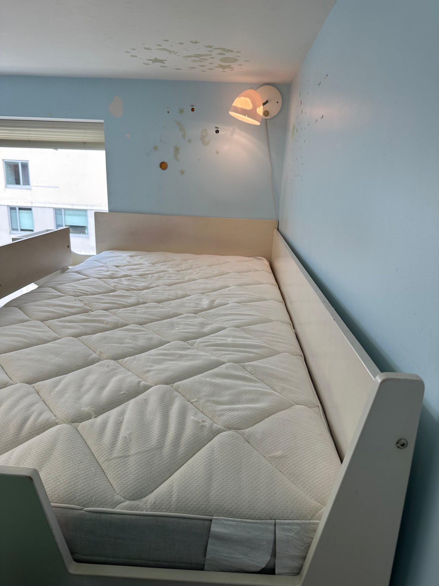 Beige bed top bunk with mattress in a room with light blue walls and a window.