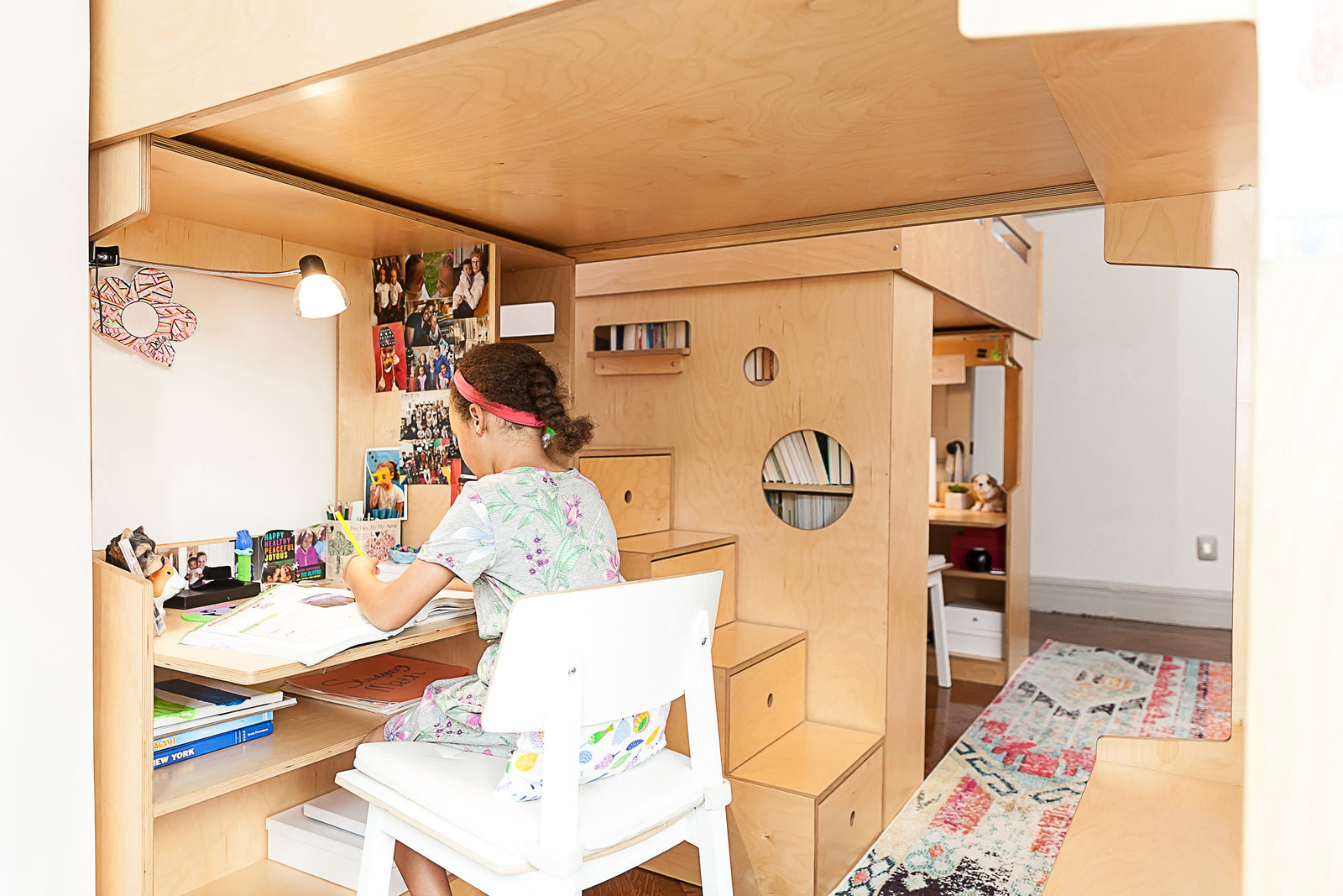 A girl sits at a wooden desk under a loft bed, working with books and a lamp, in a room with a colorful rug and cozy decor.