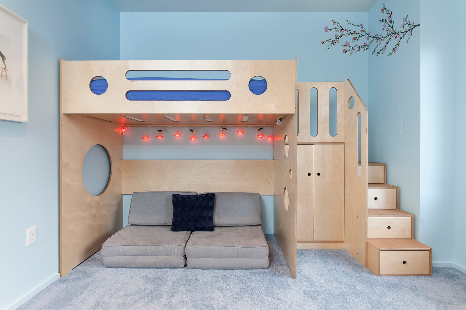 Bedroom with a light wood bunk bed, blue accents, steps with storage, gray sofa underneath, and light blue walls.