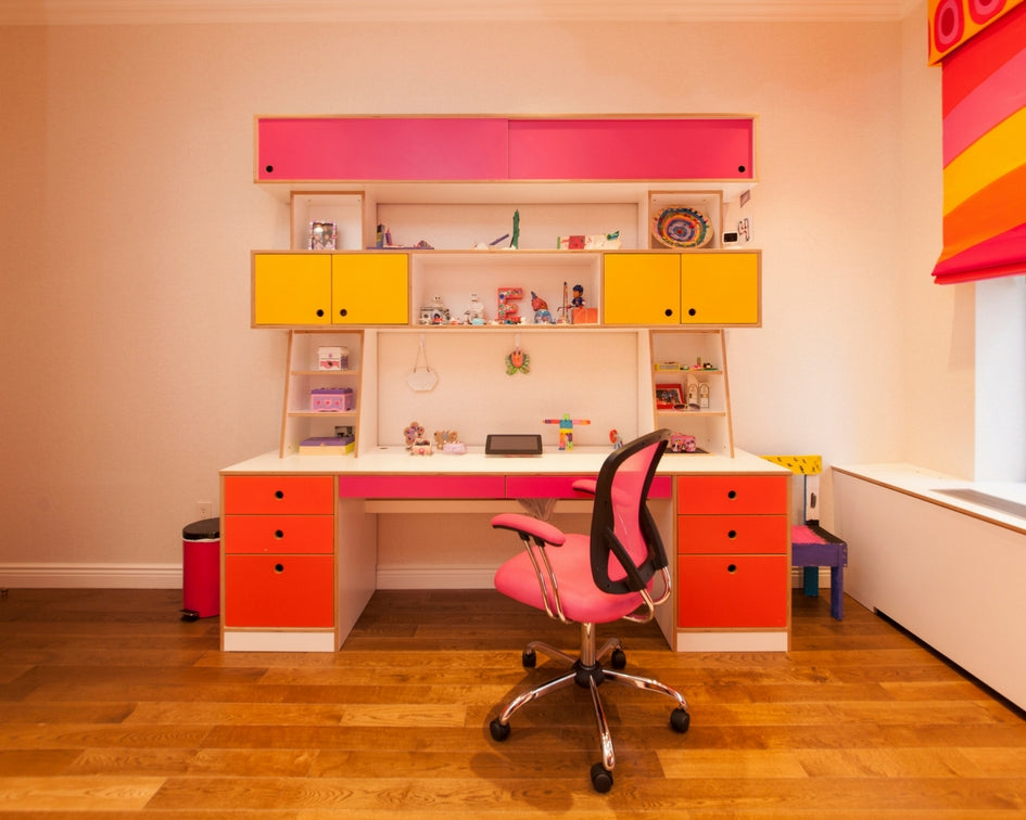 A vibrant study space with a pink and yellow desk, shelves, and a matching pink chair on a wooden floor, near a colorful window.