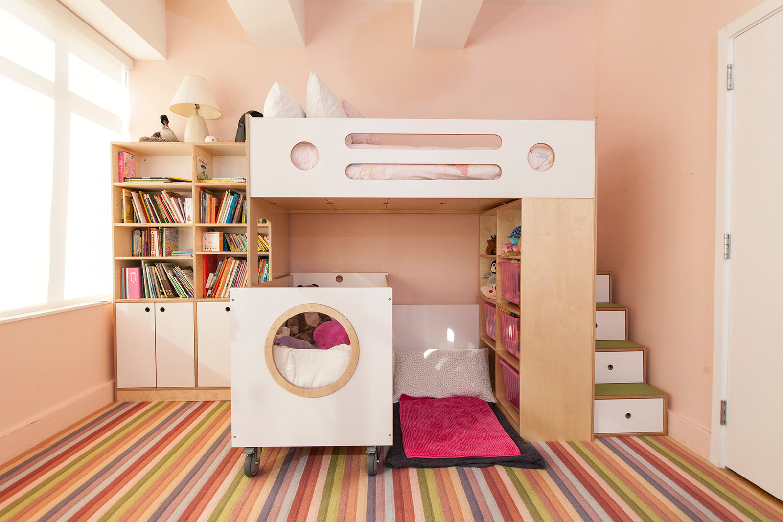 A modern bunk bed with storage stairs, a bookshelf, and a play area underneath, set in a brightly colored, striped room.