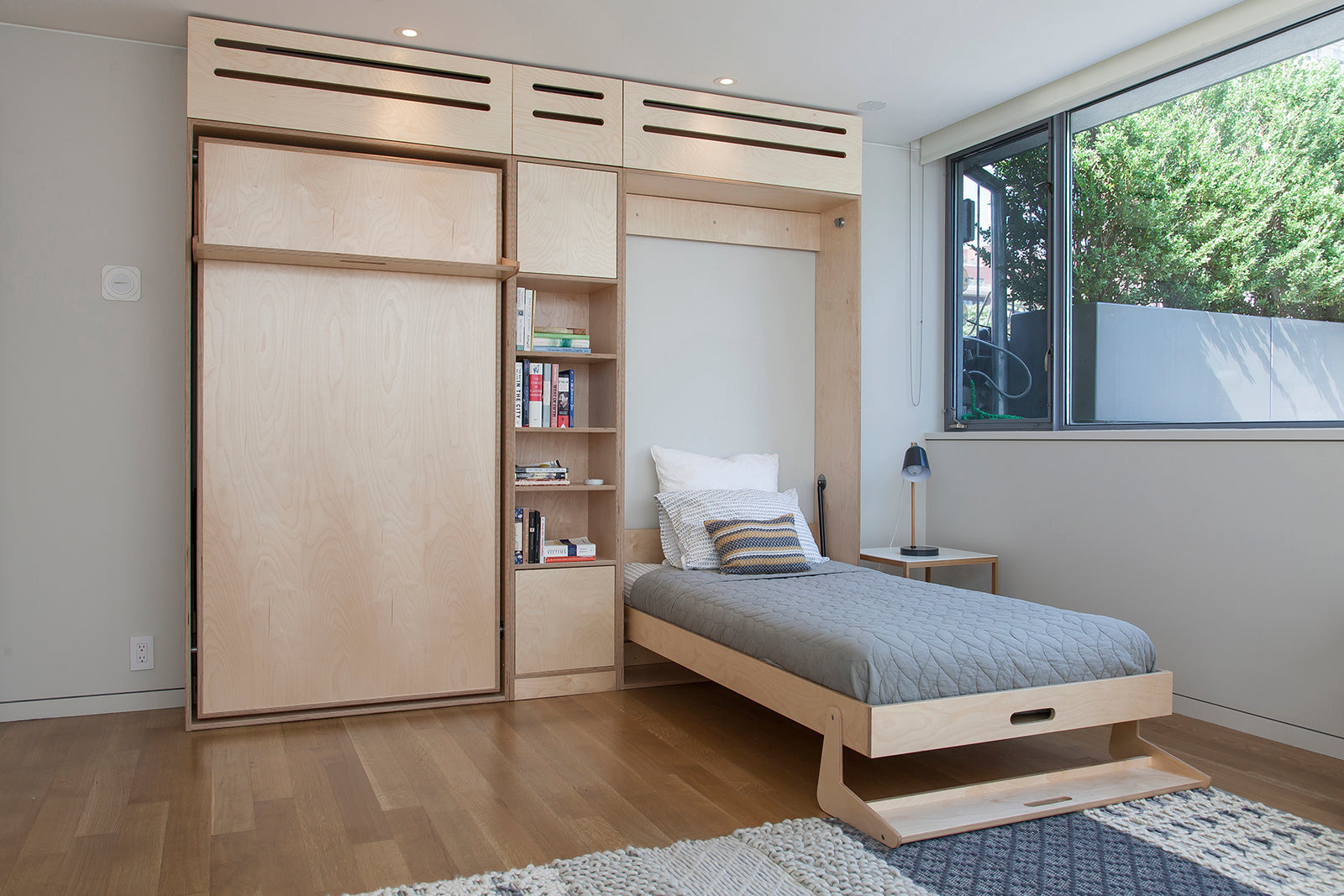 Minimalist guest room with wooden Murphy bed, built-in bookshelf, and large window with garden view.
