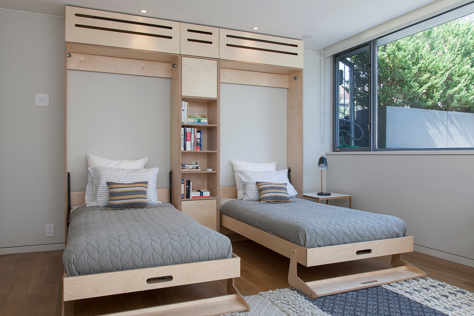 Modern room with two twin Murphy beds, wooden bookshelf, and a garden view through a large window.