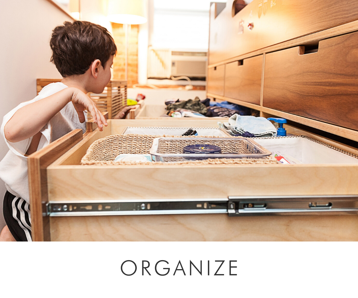Young boy organizing items in a pull-out storage drawer under a wooden bed in a well-lit room.