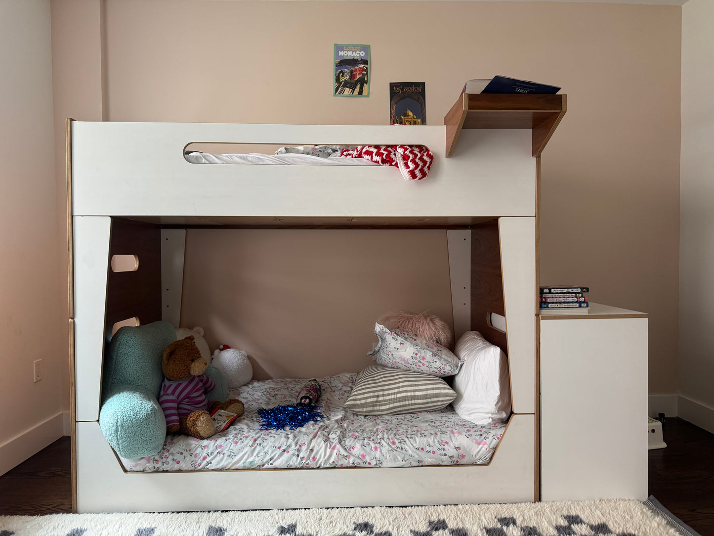 A white bunk bed frame with an open lower bed, styled with bedding, pillows, and stuffed toys. A small shelf on the right holds books, and two small framed pictures hang on the beige wall above the top bunk.