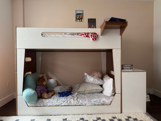 A white bunk bed frame with an open lower bed, styled with bedding, pillows, and stuffed toys. A small shelf on the right holds books, and two small framed pictures hang on the beige wall above the top bunk.