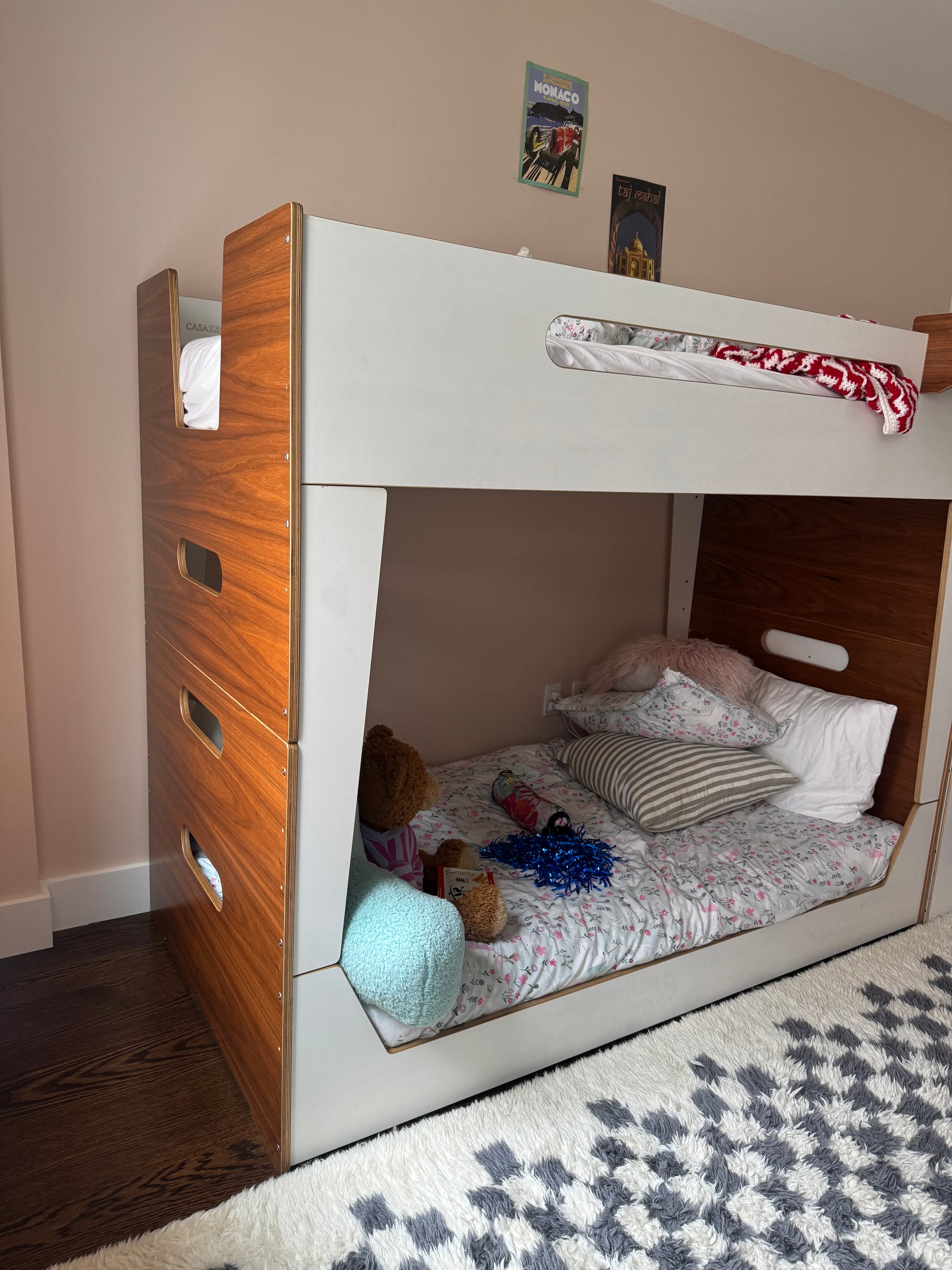 A built-in bunk bed in a child's bedroom, featuring a white frame with a warm wood-grain side drawer unit and headboard, styled with bedding and toys.