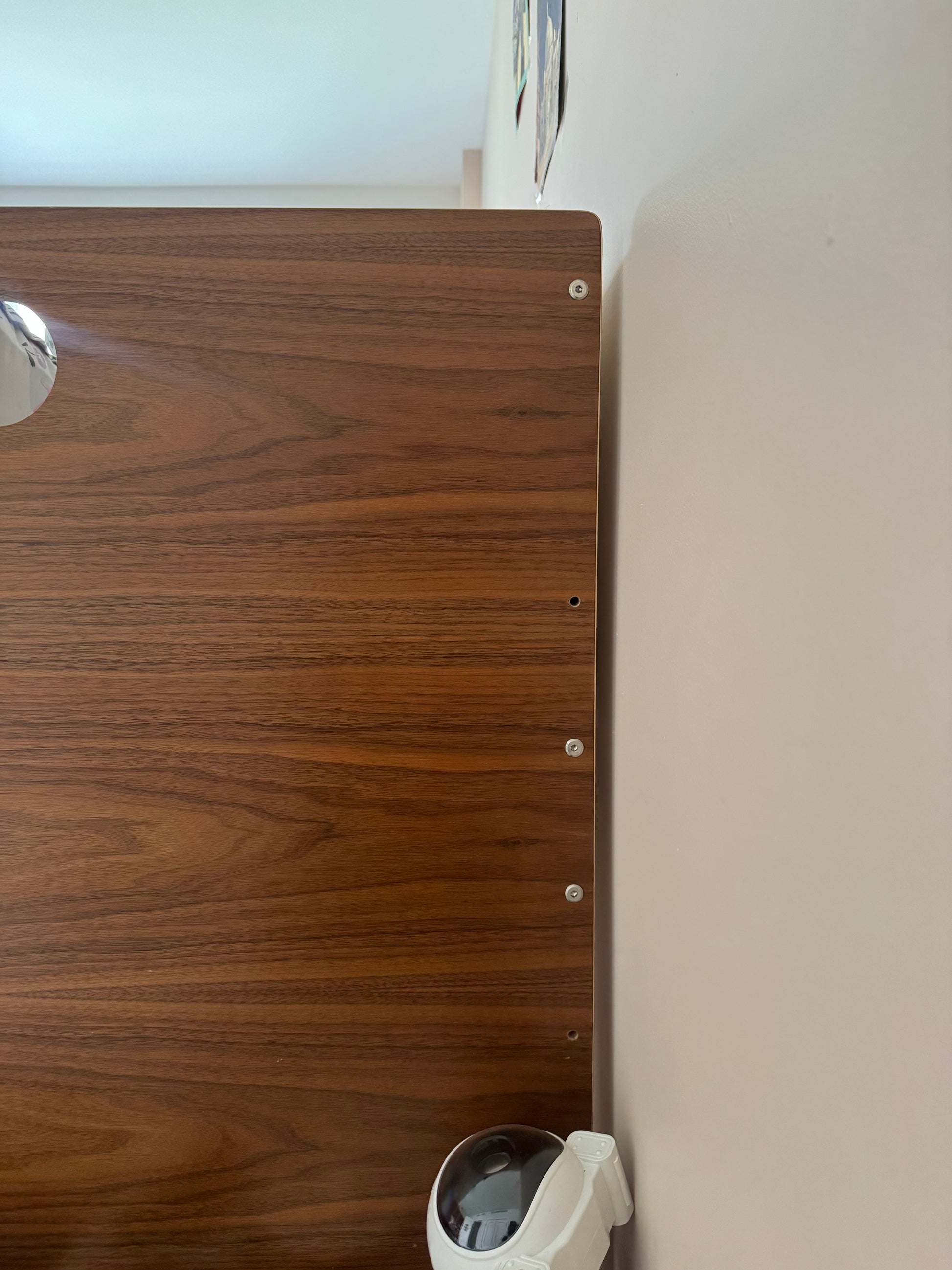A close-up of a dark brown wood grain panel with pre-drilled screw holes, mounted against a light beige wall with a white security camera visible in the lower right corner.