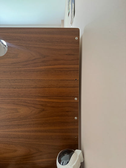 A close-up of a dark brown wood grain panel with pre-drilled screw holes, mounted against a light beige wall with a white security camera visible in the lower right corner.