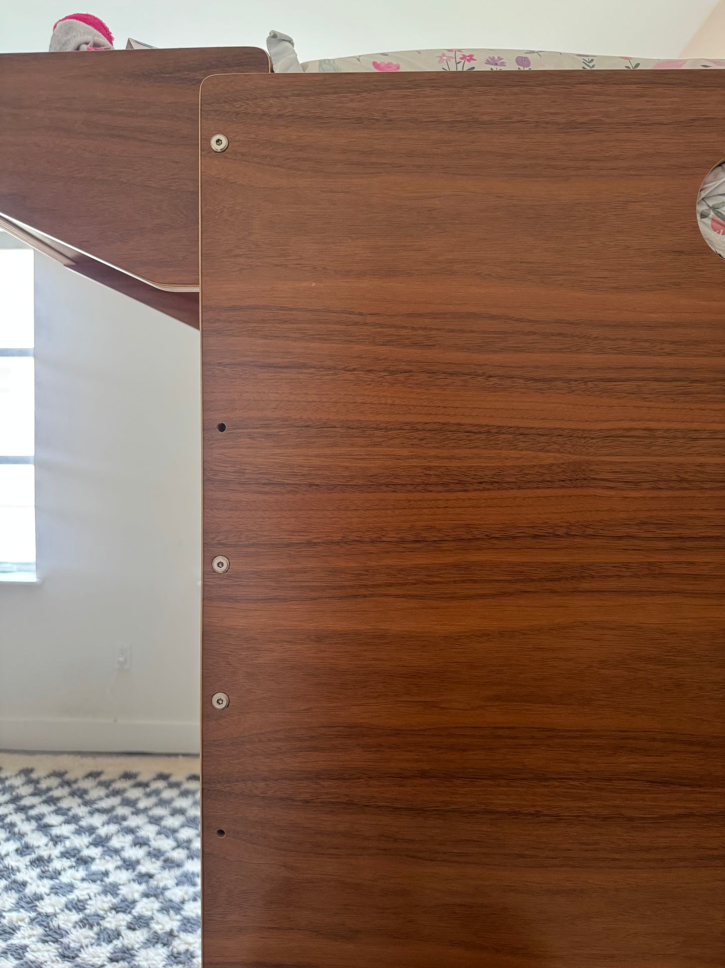 Close-up of a wooden furniture panel with a warm brown wood grain finish, showing pre-drilled screw holes and part of the furniture structure in an indoor room setting.