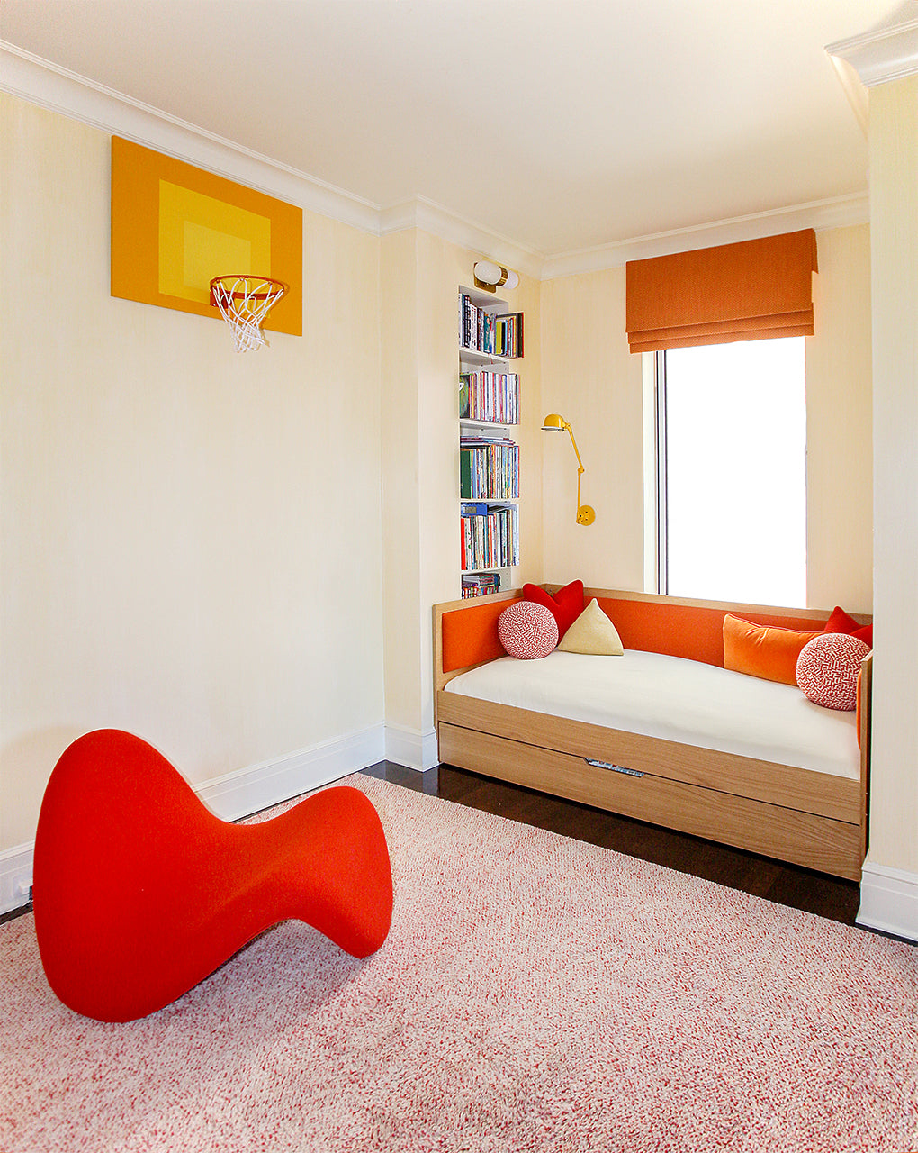 Children's room with an orange chair, basketball hoop, cozy daybed with red cushions, bookshelves, and a window with an orange blind.