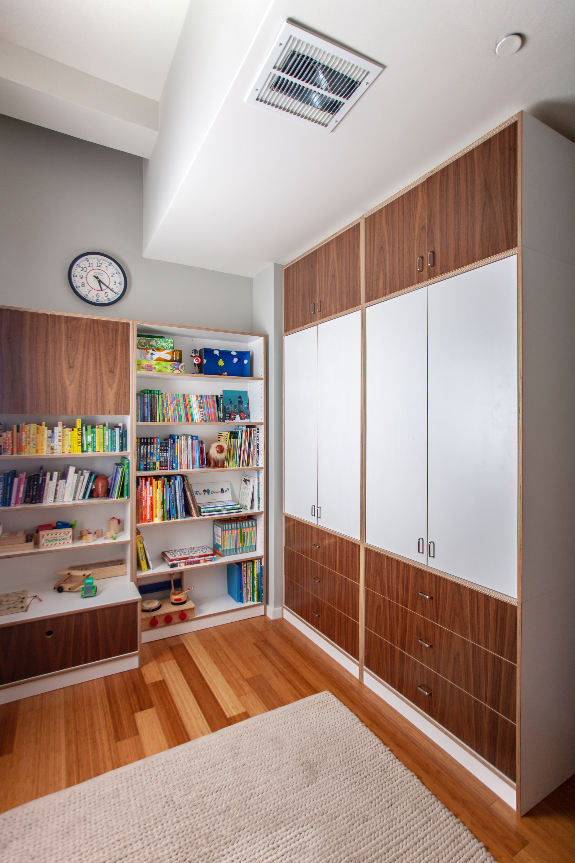 A room with wooden bookshelves filled with books and toys, a large white wardrobe, and a clock on the wall above the shelves.