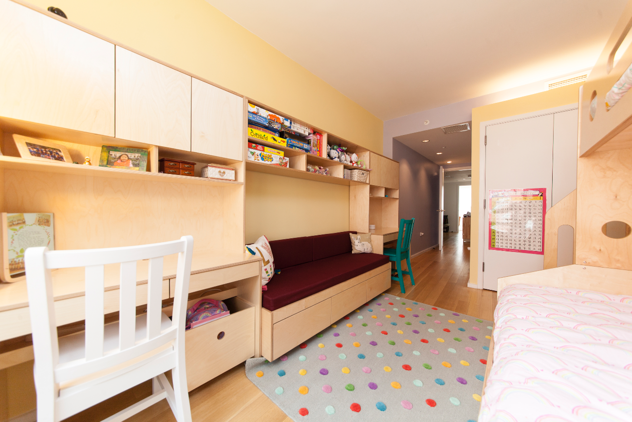 A bright, colorful kids' room with a study desk, shelves filled with toys and books, a sofa, a bed, and a polka dot rug.