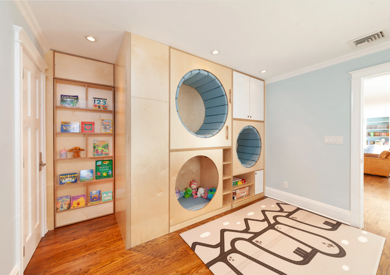 A children's playroom featuring a large light wood modular play structure with built-in bookshelves, circular tunnel nooks, and storage cubbies. The room has light wood floors, a pale blue wall, and a patterned rug on the floor.
