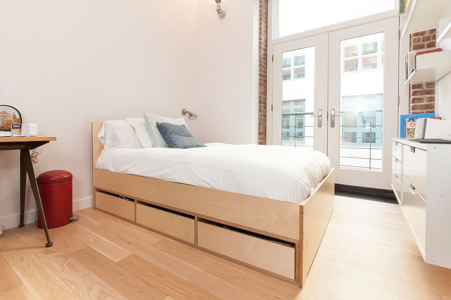 Bright bedroom with a wooden bed, white bedding, desk, red bin, and large French doors opening to a balcony.