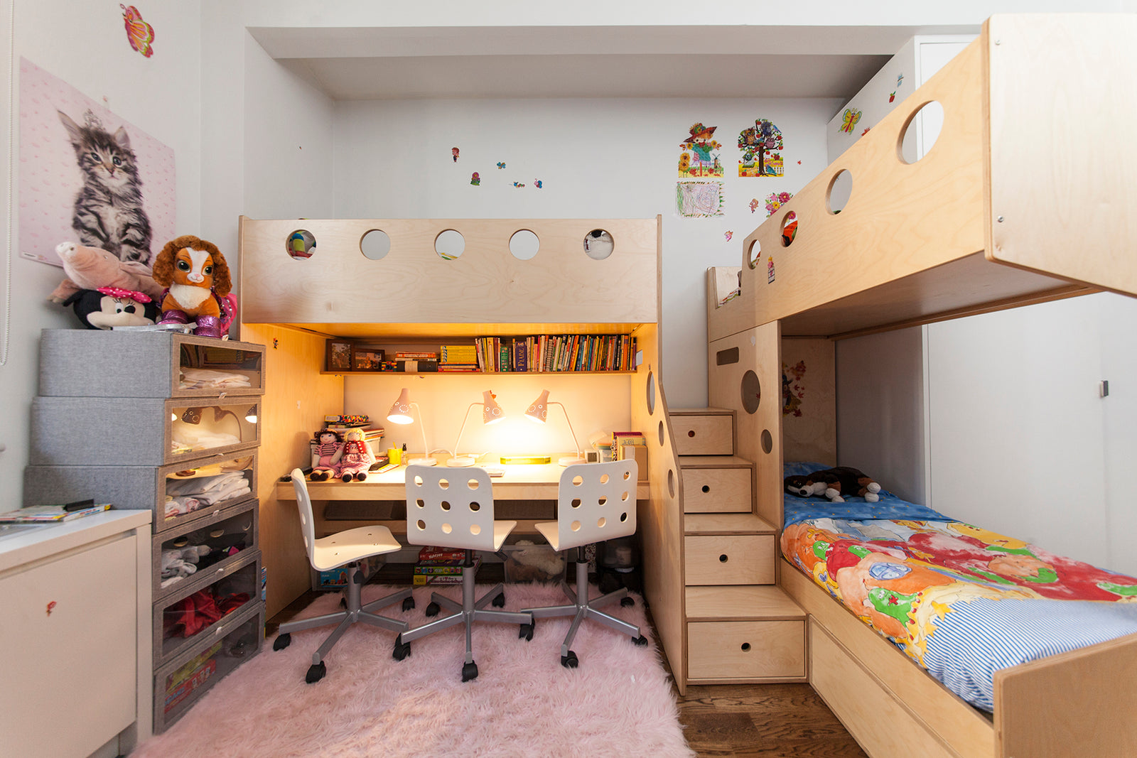 A shared kids' room with a wooden bunk bed, study area with two chairs, colorful bedding, and plush toys on shelves.