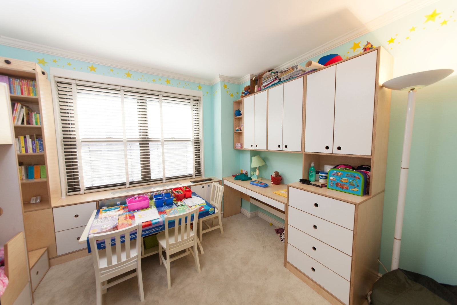 A custom-built birch and white storage and desk unit in a children's learning space, featuring upper cabinets, drawers, open shelving, and a built-in workspace. A large window seat with additional storage below provides extra functionality, while a colorful play table is set up in the center of the room