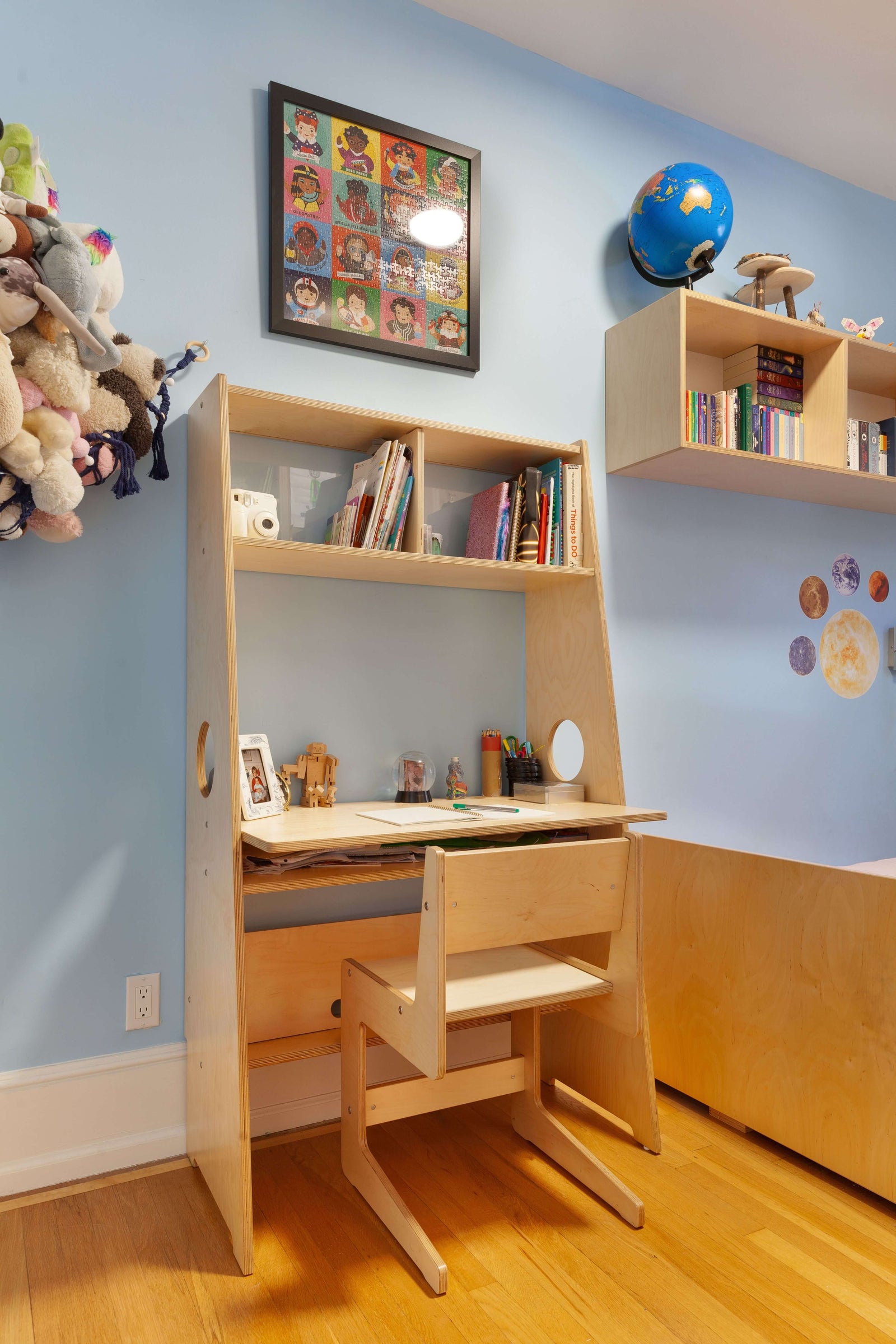 A compact birch-finish desk with built-in shelving, paired with a matching adjustable chair. The workspace includes open storage for books and supplies, with a clean and functional design. A floating bookshelf and decorative elements, including a globe and space-themed wall decals, add personality to the room.