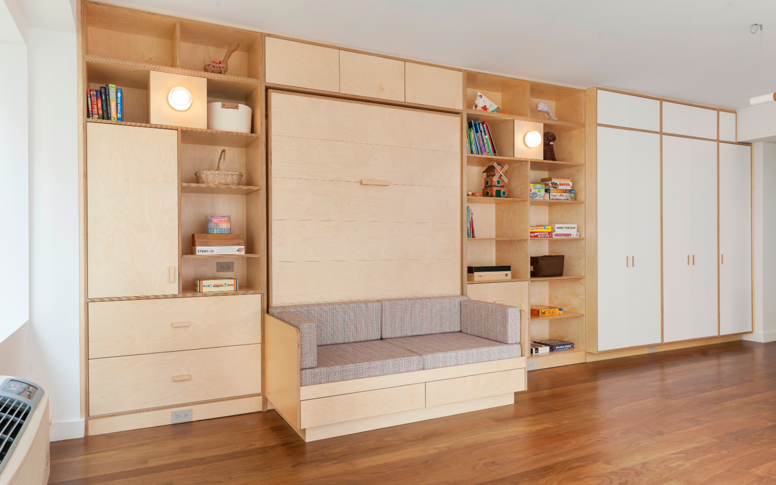 Custom-designed birch Murphy bed wall unit with integrated shelving, cabinets, and a built-in sofa. The sofa sits below the Murphy bed, which can be pulled down to create a guest sleeping area. The unit maximizes storage and functionality in a bright, organized space.

