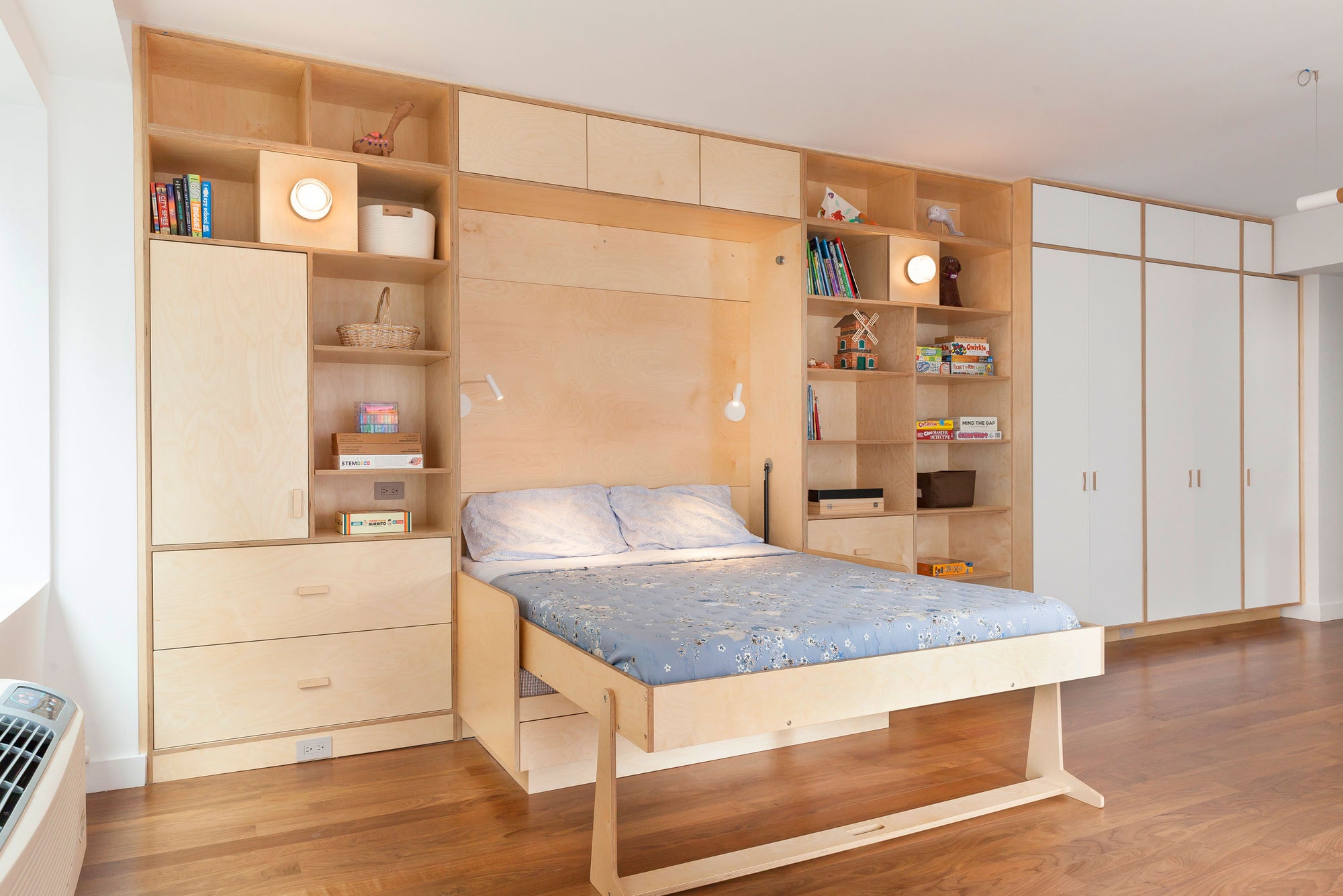 Spacious room with a custom Murphy bed integrated into a birch storage wall, featuring open shelves, cabinets, and drawers. The setup provides a functional sleeping area that can be folded away, maximizing space with ample storage for books, toys, and decor.

