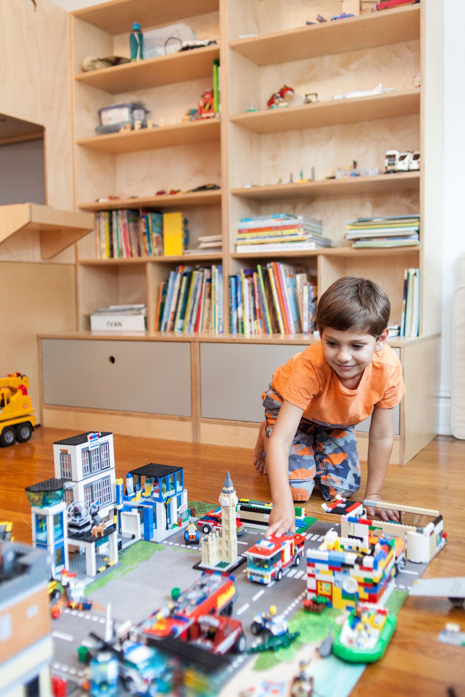 A custom birch storage cabinet with open shelving and lower drawers provides ample organization for books, toys, and LEGO creations. A young boy enjoys playing with an elaborate LEGO city setup on the floor, showcasing a playful and functional children's space.