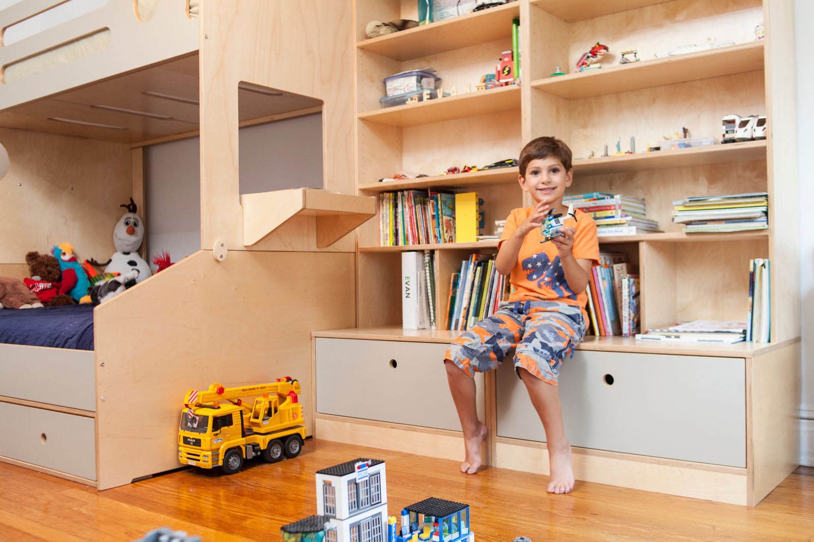 A modern twin-over-full bunk bed in natural birch with gray accents, integrated with custom storage cabinets featuring drawers and open shelving. The space is designed for organization and play, with books, toys, and LEGO creations neatly displayed. A young boy sits on the built-in storage bench, holding a LEGO helicopter, adding a warm, playful atmosphere to the room.