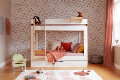 Birch and white Cabin Bunk Bed with rectangular cutouts, attached ladder, a trundle bed, and grey bedding against rainbow print wallpaper on a wood floor