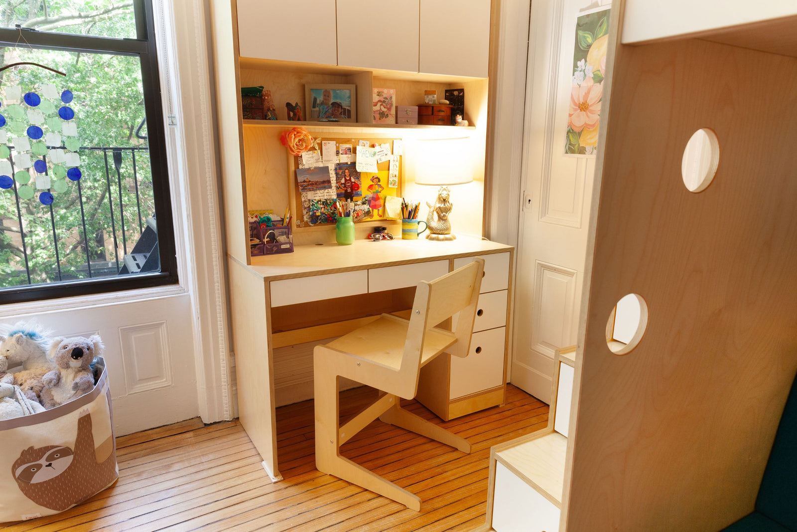 A custom birch and white desk with built-in storage drawers and a matching adjustable chair. The workspace features upper shelving, a soft-lit lamp, and a corkboard with personal mementos. Positioned near a window with natural light, the desk creates a cozy and functional study area, complemented by a staircase with integrated storage leading to a lofted bed.