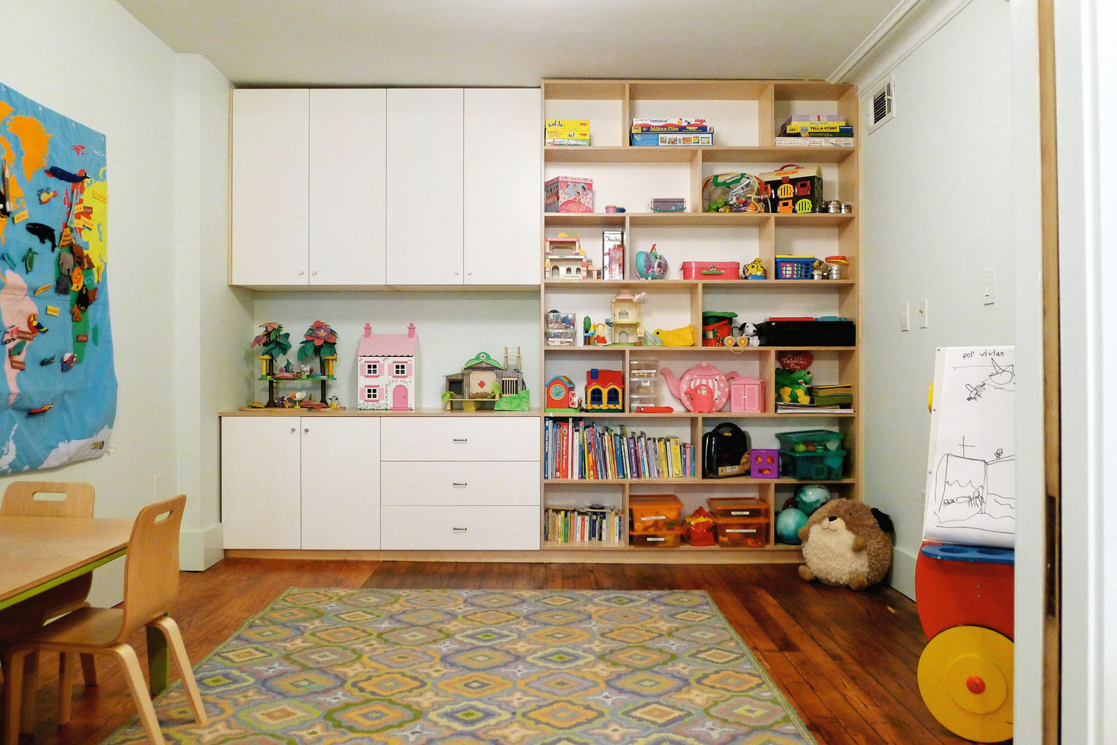 A custom birch and white storage unit featuring upper cabinets with doors, lower drawers, and open shelving filled with books, toys, and educational materials. The well-organized space includes a colorful play area with a rug, wooden chairs, and a felt world map on the wall, creating a functional and inviting environment for children