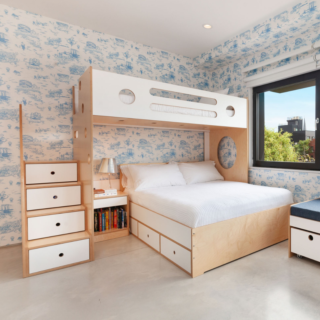 Custom bunk bed setup in birch and white finish, featuring a lofted twin bed above a full-sized bed. The design includes a staircase with built-in storage drawers on the left side, offering easy access to the top bunk. The bed is positioned against a wall with whimsical blue-and-white wallpaper, creating a playful atmosphere. Beneath the lower bed, additional drawers provide extra storage space. 