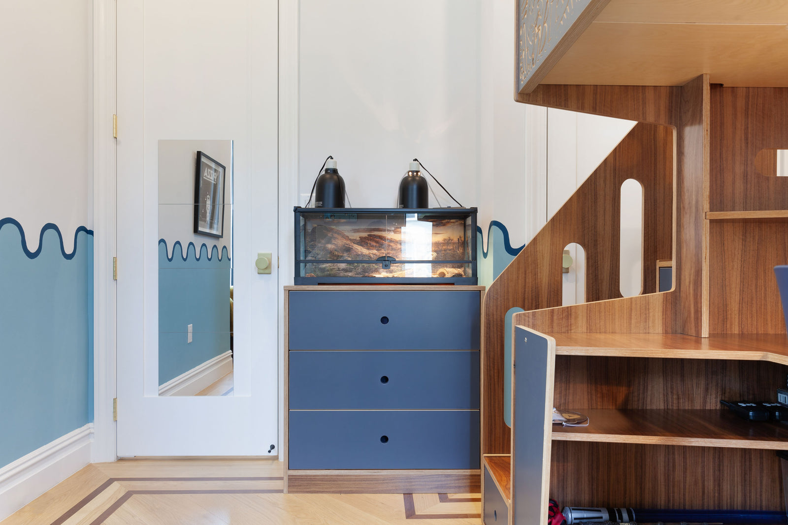 Blue dresser with a reptile tank on top, next to a walnut staircase leading to a loft bed in a children's room with blue wave-themed walls.

