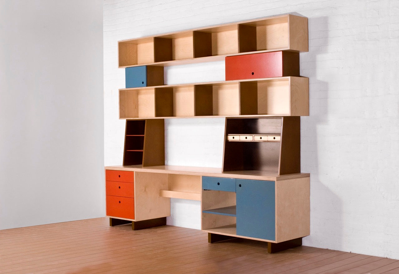 A modern wooden desk and shelving unit with colorful drawer fronts in red, blue, and black, set against a white wall.