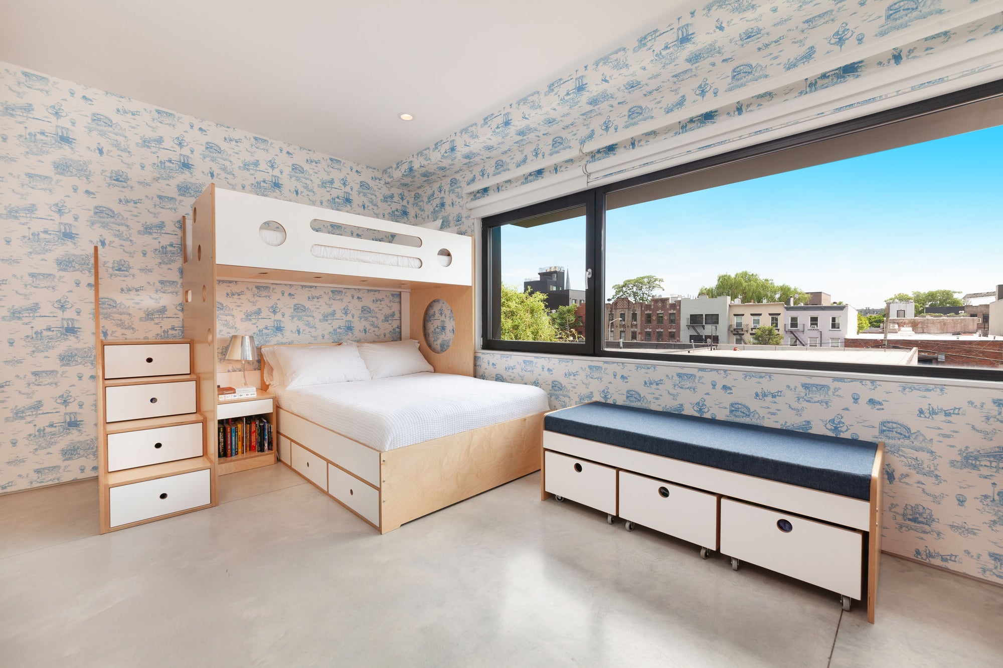 Children's bedroom with a custom birch bunk bed featuring white accents, built-in storage drawers, and a staircase with drawer steps. The room includes a window bench with storage drawers on wheels and blue upholstery, providing additional seating and storage in a bright, playful space.

