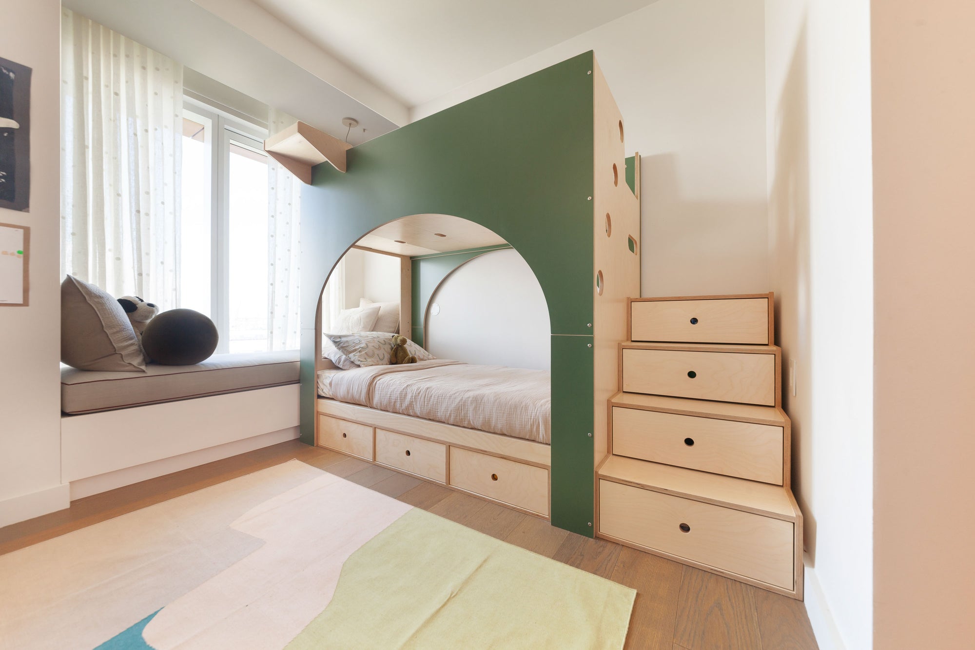 Custom kids' bunk bed with a distinctive green arch design, birch finish, and integrated storage drawers beneath the bed and in the staircase. The cozy setup includes a built-in window bench, creating a playful and functional sleeping area that maximizes space.

