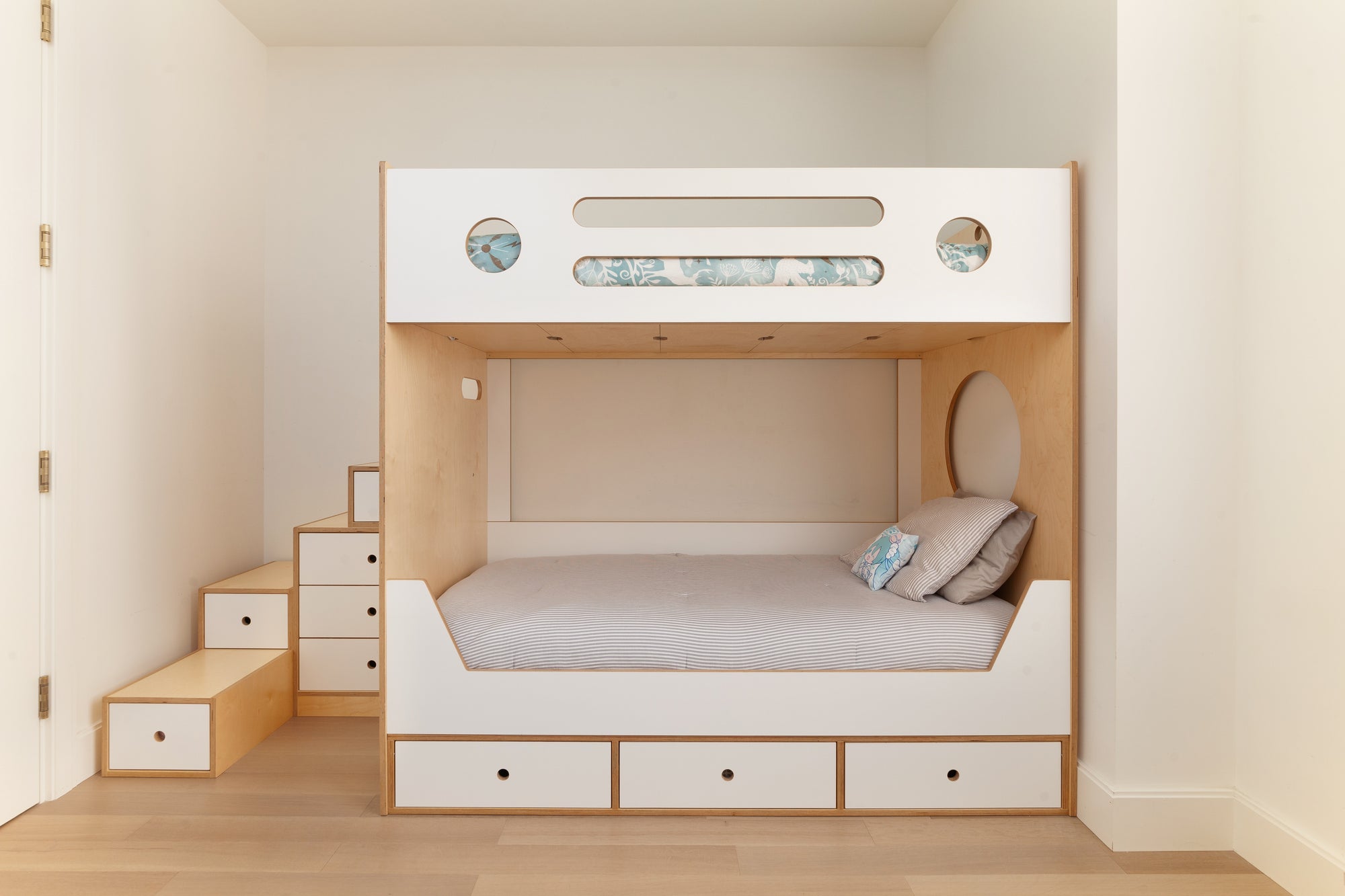 Custom bunk bed in a birch and white finish, featuring integrated storage drawers under the lower bunk and in the staircase. The design includes circular cutouts on the upper bunk and cozy bedding, creating a practical and stylish sleeping area for kids.

