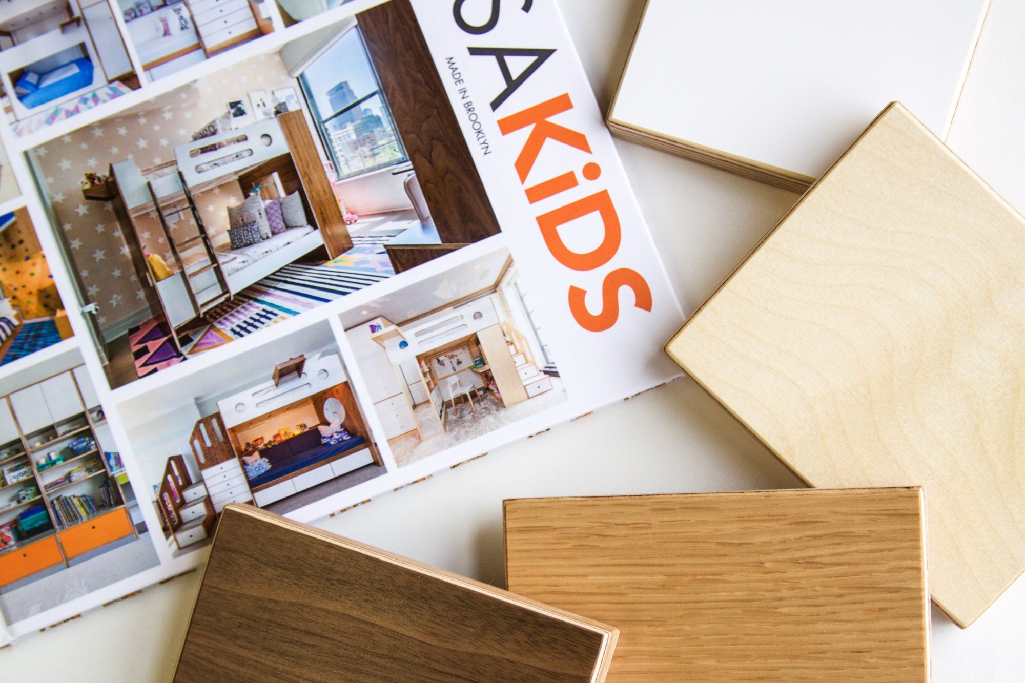 A close-up of the Casa Kids portfolio book, showcasing custom-built bunk beds and storage solutions, alongside four main wood finish samples. The finishes include natural birch, white laminate, warm oak, and rich walnut, representing the brand’s signature aesthetic. The image highlights the craftsmanship and design versatility of Casa Kids furniture.