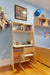 Children's desk setup with built-in shelf, light wood finish against a blue wall. Desk is neatly organized with books, globe, and art decor, creating a cozy study nook.

