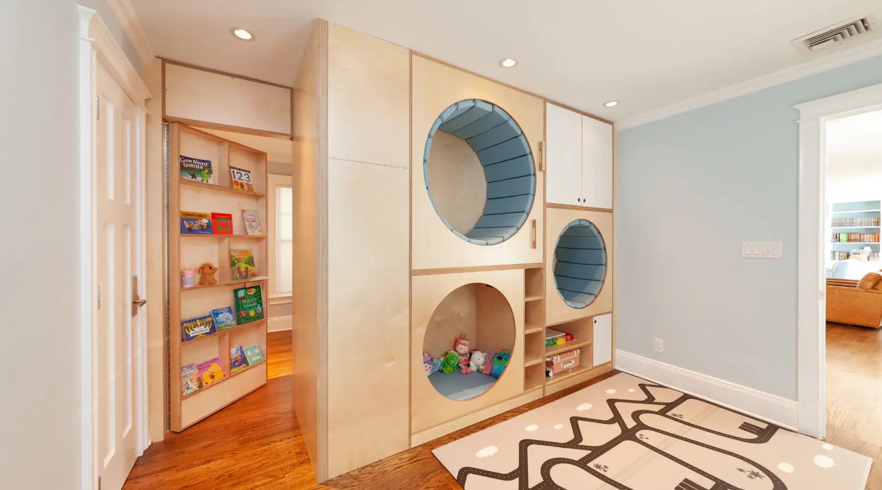 Custom birchwood playroom storage unit with built-in circular nooks lined with blue cushions, toy cubbies, cabinets, and a hidden bookcase door leading to an adjacent space