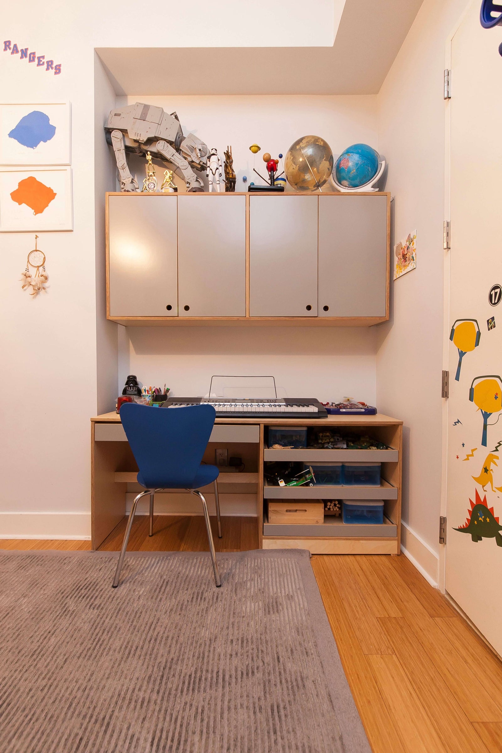 A custom-built desk with drawers and an upper storage cabinet, designed for a compact workspace. The desk features open shelving for organization, a built-in keyboard area, and a blue accent chair. The upper storage cabinets provide additional space for books, toys, and collectibles, including LEGO sets and globes. A playful, creative atmosphere is enhanced by wall decorations and artwork.