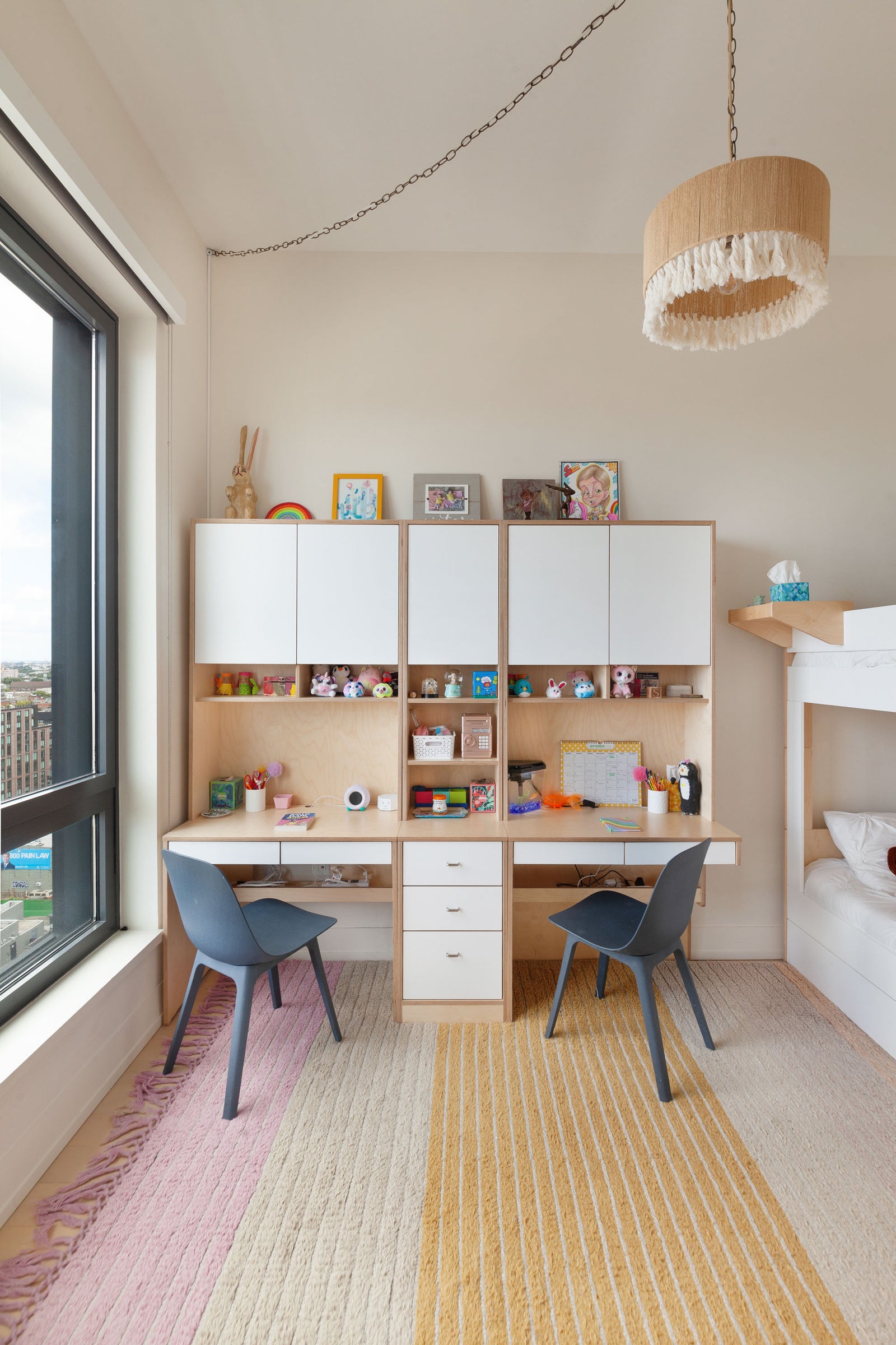 Custom-built shared kids' study area featuring a spacious double workstation with a natural birch finish, white overhead cabinetry, and open shelving. The desk setup includes two black chairs, ample drawer storage, and personalized decor. A white bunk bed is partially visible on the right, and a large window on the left provides natural light